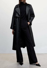 Trench-coat en cuir noir avec grands revers, fermeture à boutons, associé à un col roulé noir ajusté et un pantalon noir taille haute.