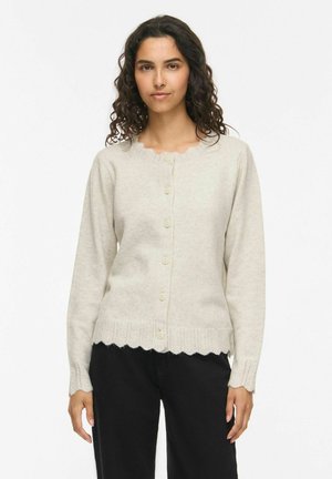 Jeune femme aux cheveux bouclés portant un cardigan en maille beige boutonné avec des bords festonnés et un pantalon noir, debout devant un fond uni.