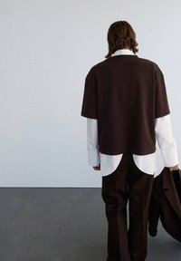 Brun oversized top båret over en hvid skjorte med buet kant, parret med mørke bukser. Glat stof, løs pasform og lagdelt design.