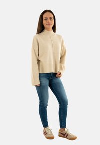 Pull en maille beige avec un col montant et un ourlet à côtes, associé à un jean bleu et des baskets beiges avec des accents bordeaux.