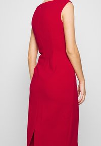 Robe sans manches rouge avec une silhouette ajustée, dotée d'une fente dans le dos et d'un détail de fermeture éclair discret au niveau du col. Texture de tissu lisse.