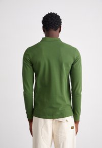 Camisa polo de manga longa em algodão verde escuro com colarinho e punhos canelados, usada com calças bejes claras com bolsos e uma etiqueta.