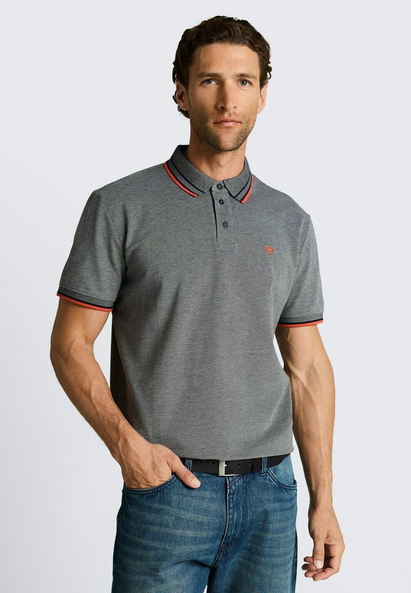 Hombre con una camiseta polo gris ajustada con cuello y bordes de las mangas a rayas rojas y negras, combinada con vaqueros azules y un cinturón negro.