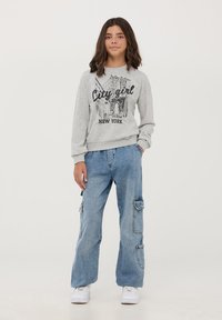 Szara bluza z nadrukiem "City Girl New York", zestawiona z jasnoniebieskimi spodniami cargo z kieszeniami i luźnym fasonem. Białe sneakersy.