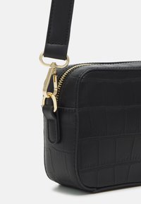 Černá taška z umělé kůže s krokodýlím vzorem, se zlatým zipem, kovanými detaily a plochým popruhem. Kompaktní obdélníkový tvar.