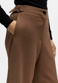 Asmuo vilki aukšto liemens rudas kelnes su šonine kišene ir sagtimi, derindamas su juodu ilgomis rankovėmis marškiniais.