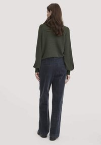 Groene gebreide trui met ruime mouwen, gecombineerd met marineblauwe corduroybroek met een ontspannen pasvorm en subtiele textuur. Zwarte schoenen maken de look compleet.