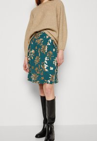 Tunika im Türkis mit floralen Motiven und metallisch goldenen Akzenten, kombiniert mit einem beigen, gerippten Pullover und schwarzen kniehohen Stiefeln. Sanfte Textur und maßgeschneiderte Passform.