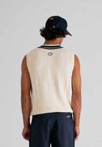 Man wearing cream cable-knit sleeveless sweater with navy trim and navy pants, facing away against a plain grey background.