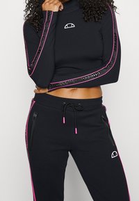 Mustat lyhyt toppi vaaleanpunaisilla yksityiskohdilla ja brändäyksellä hihoissa, yhdistettynä mustiin collegehousuihin, joissa on vetoketjulliset taskut ja vaaleanpunaiset sivuviivat.