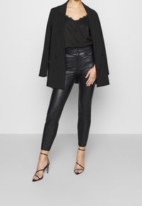 Svart blazer över en cami med spetsdetaljer, paired med högmidjade svarta byxor i konstläder och svarta remsandaler. Slät textur, figurnära design.