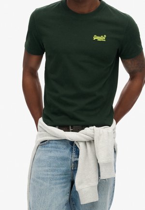 Dark green cotton T-shirt with a round neck and small yellow logo on the chest. Paired with light blue jeans and a gray sweatshirt tied at the waist.