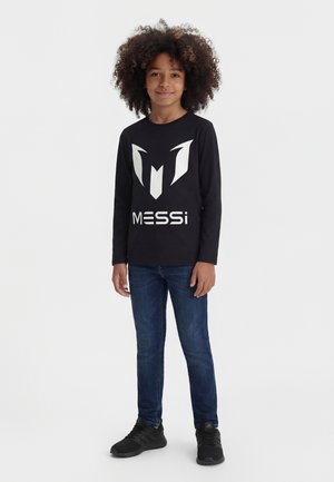 Kind mit lockigem Haar, trägt ein schwarzes Langarmshirt mit Messi-Motiv, blaue Jeans und schwarze Turnschuhe, steht vor einem schlichten weißen Hintergrund.