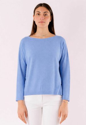 Giovane donna con lunghi capelli scuri che indossa un maglione a maniche lunghe azzurro chiaro e pantaloni bianchi su uno sfondo uniforme.