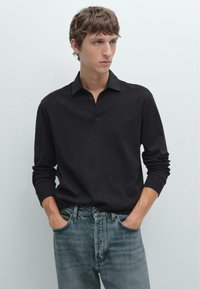 Schwarzes langärmliges Polohemd mit Kragen und Knopfleiste. Trägt dazu eine hellblaue Jeans, die einen lässigen Look zeigt.