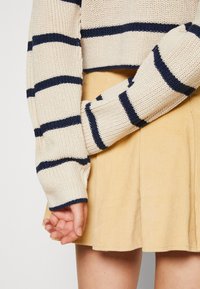 Pull en tricot crème avec des rayures bleu marine sur les manches, associé à une jupe en velours côtelé jaune, mettant en avant la texture et les couleurs contrastées.