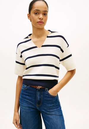 Kurzärmeliger Pullover mit V-Ausschnitt in Creme mit marineblauen horizontalen Streifen. Lockerer Schnitt mit geripptem Saum, kombiniert mit blauen Jeans und einem braunen Gürtel.