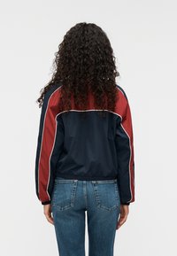 Άνεμος με χρώματα ναυτικού μπλε και βουργουνδί με λευκές λεπτομέρειες. Διαθέτει σφιχτή ζώνη στη μέση, μακριά μανίκια και άνετη εφαρμογή. Το σύνολο ολοκληρώνουν τζιν παντελόνια.