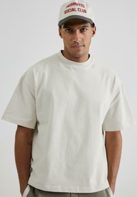 Tricou alb din bumbac, cu umeri lăsați și guler rotund. Modelul poartă o șapcă gri cu inscripția "CLUB SOCIAL INTERZIS" în text roșu.