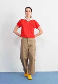 Red short-sleeve sports top with white collar and yellow accents, paired with beige cargo pants featuring side pockets and yellow shoes.