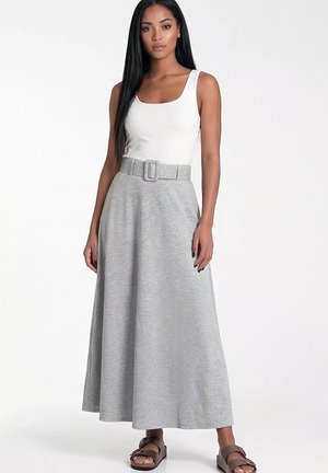 Kobieta z długimi czarnymi włosami, ubrana w biały bezrękawnik, szarą maksisukienkę z paskiem i brązowe sandały na wsunięcie, stojąca na jednolitym tle.