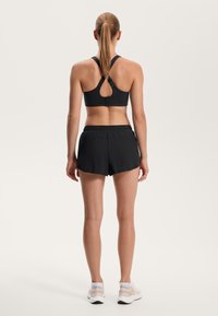 Čierny športový podprsenka s otvoreným chrbtom a prekríženými ramienkami v kombinácii s čiernymi bežeckými šortkami. Viditeľná hladká textúra a elastický pás.