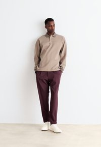 Beige long-sleeve polo shirt with a subtle collar detail, paired with burgundy trousers and white sneakers, set against a neutral background.