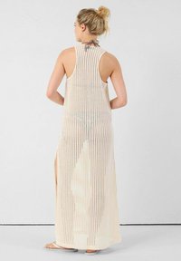 Lange, mouwloze cover-up jurk van lichtgewicht, beige gebreid materiaal, met een gaaspatroon en zijsplitten voor luchtcirculatie en bewegingsgemak.