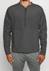 Pull-over gris foncé à manches longues avec un col à demi-fermeture éclair, doté d'un tissu lisse, de coutures contrastantes et de poches latérales.