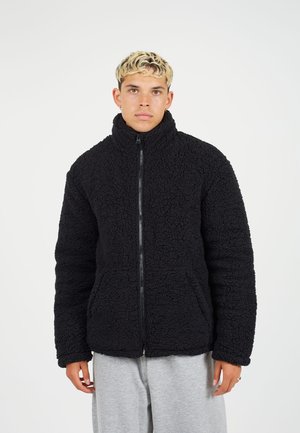 Chaqueta de forro polar negra con una superficie texturizada, cuello alto, cremallera completa y bolsillos laterales. Presenta un ajuste holgado y una tela suave.