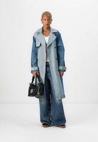 Denim trench coat with two-tone blue shades, oversized fit, large collar, and double-breasted buttons. Paired with wide-leg jeans and black handbag.