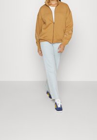 Haut de survêtement zippé marron avec un col haut, associé à un jean droit bleu clair et des baskets bleues avec des rayures jaunes.