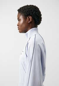 Chaqueta de cremallera azul claro con un alto cuello, que presenta ribetes oscuros en contraste y una tela texturizada. Un bolsillo lateral visible.