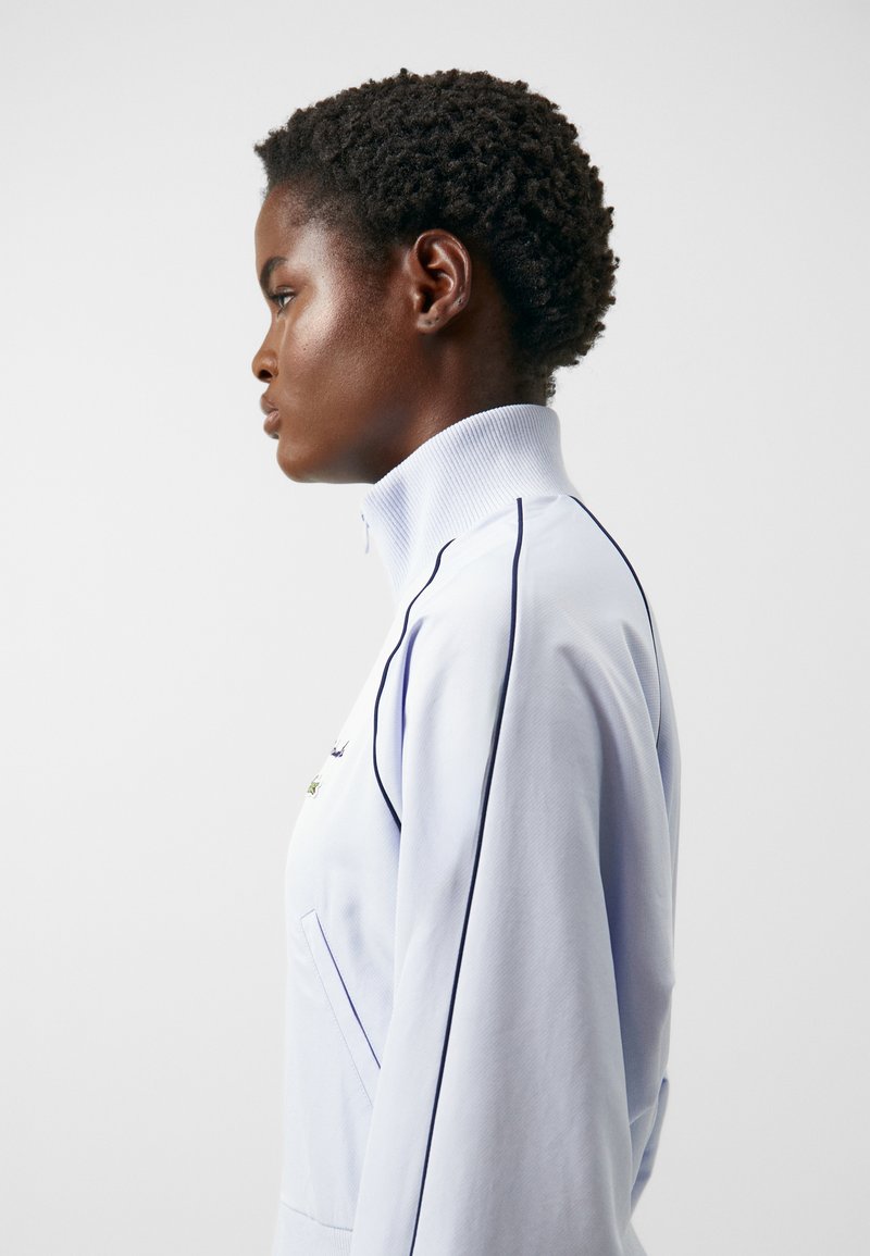 Hellblaues Zip-Jackett mit hohem Kragen, das kontrastierende dunkle Paspeln und einen strukturierten Stoff aufweist. Seitentasche sichtbar.