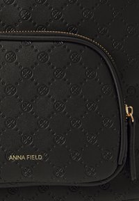 Sac en cuir noir texturé avec une poche zippée à l'avant ornée de motifs floraux en relief. Détail de marque doré portant l'inscription "ANNA FIELD."