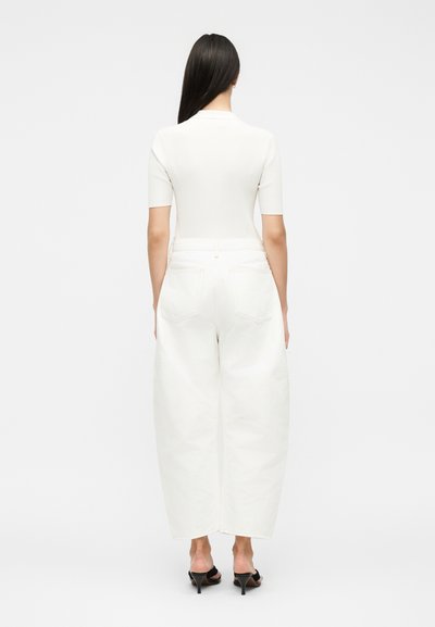 Γυναίκα με μακριά σκούρα μαλλιά, φορεί εφαρμοστό λευκό μπλουζάκι με κοντά μανίκια, φαρδιά λευκά παντελόνια και μαύρα σανδάλια με τακούνι, στέκεται με την πλάτη στην κάμερα.