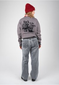 Femme portant un bonnet rouge en tricot, un sweat-shirt gris avec "Back in the Days" et un graphisme de véhicule, ainsi qu'un jean gris délavé, debout de dos.