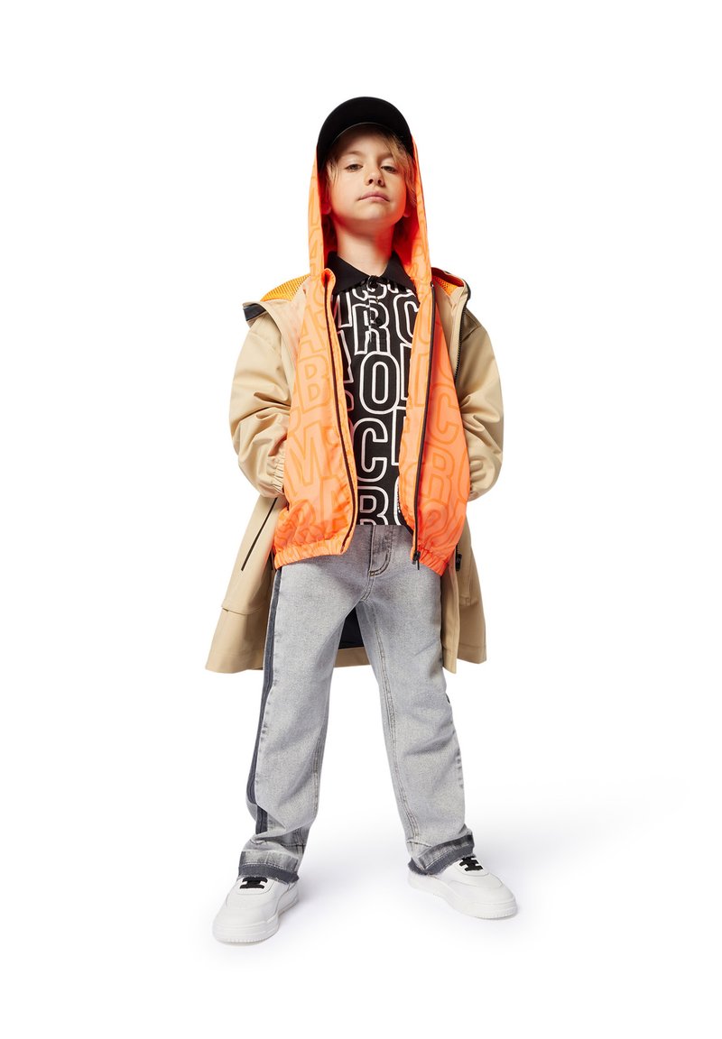 Kind dat een beige jas, een fel oranje capuchonjack, een zwart shirt met opdruk, lichte spijkerbroek en witte sneakers draagt.