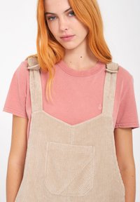 Beige kordfløjl overaller med en frontlomme og justerbare stropper, båret over en pink crew-neck t-shirt. Glat tekstur, afslappet design.