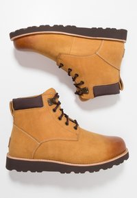Botas de tobillo de cuero marrón claro con cordones, con un cuello acolchado marrón oscuro y suelas negras robustas, mostradas una al lado de la otra sobre un fondo blanco.