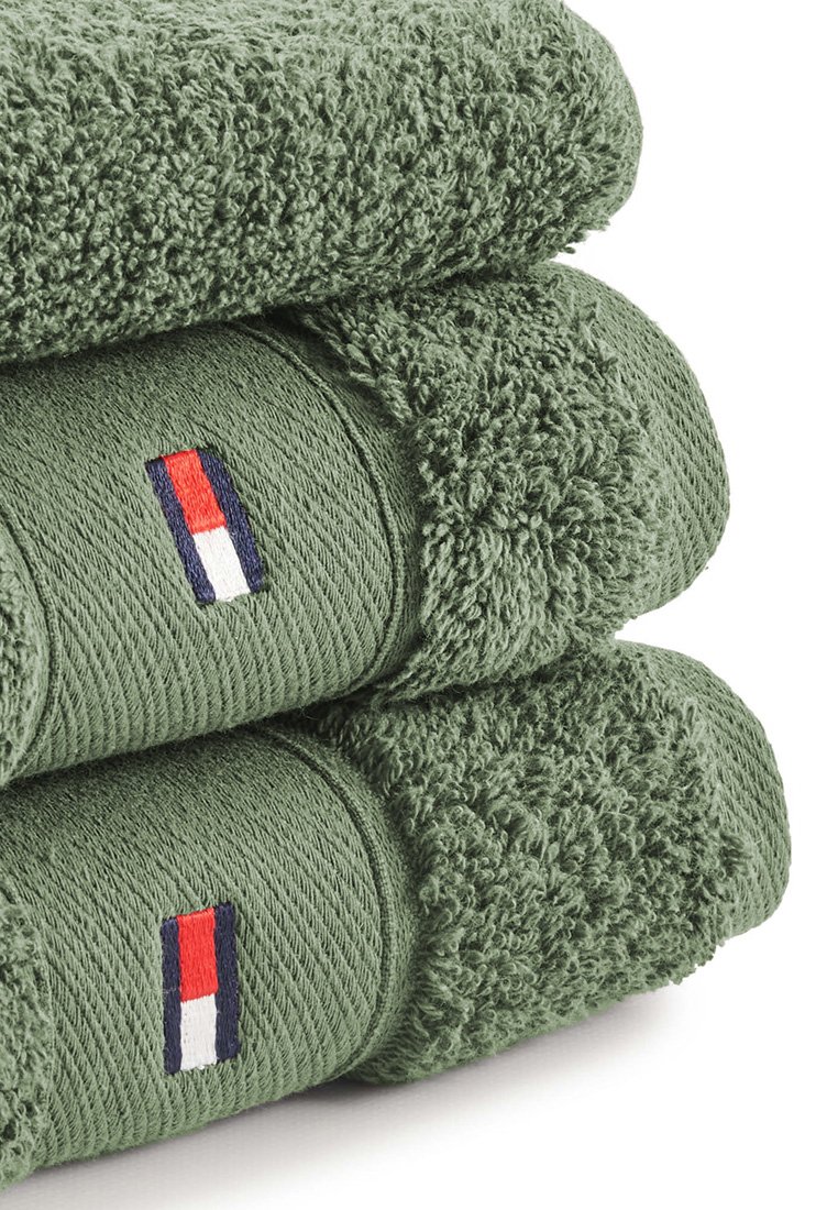 Zieleń ręczników frotte ułożonych w stertę, z żebrowanymi i teksturowanymi powierzchniami. Każdy ręcznik ma małe haftowane logo w akcentach czerwonym, białym i granatowym.