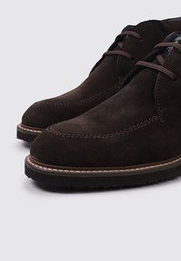 Zapatos de ante marrón oscuro con punta redondeada, detalles de costura a lo largo de la parte superior y una suela de goma con un contorno de costura clara en contraste.