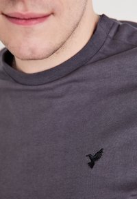 Γκρι βαμβακερό μπλουζάκι με στρογγυλή λαιμόκοψη και μικρό μαύρο κεντημένο λογότυπο πουλιού στο στήθος. Μαλακή υφή και χαλαρός σχεδιασμός.