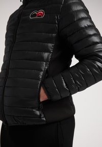 La giacca a vento nera presenta un design trapuntato con una texture lucida, tasche con zip e un pannello laterale in tessuto scuro a contrasto.