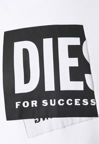 Bílý bavlněný tričko s černým obdélníkovým nášivkem, na kterém je velkým bílým písmem nápis: "DIES FOR SUCCESS" na přední straně. Hladká textura.