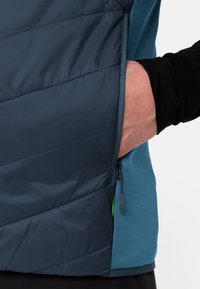 Gilet isolato blu navy con cuciture diagonali, tasca laterale e dettaglio con zip. Presenta un tessuto di contrasto blu chiaro sui lati.