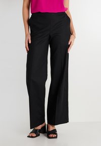 Pantalons larges noirs avec une texture lisse, présentant des fentes sur les côtés et une taille plate. Associés à des sandales à talons noires et un haut rose.