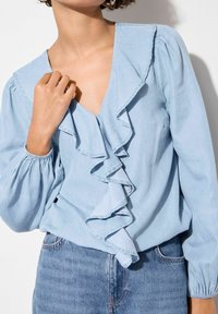 Blusa azul clara con escote en V profundo, con un frente con volantes y mangas largas abullonadas. Combinada con jeans de mezclilla azul de cintura alta.