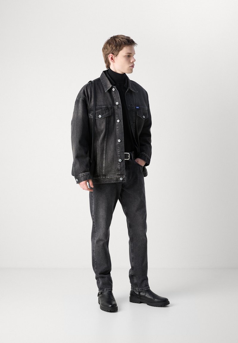 Czarna dżinsowa kurtka z dużymi kieszeniami i guzikami, zestawiona z dopasowanymi czarnymi dżinsami i czarnymi botkami za kostkę. Gładka faktura, minimalistyczny design.