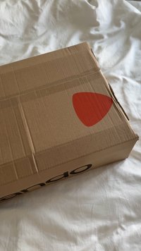 Rjava kartonska škatla s trikotno rdečo nalepko na vrhu in črnim napisom ob strani. Škatla je videti zapečatena s trakcem in ima teksturirano površino.